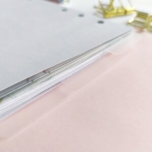 May include: A stack of white paper sheets with punched holes along the left edge, featuring translucent plastic index tabs extending from the right side. Two gold-toned metal binder clips are positioned near the top right corner of the paper stack.