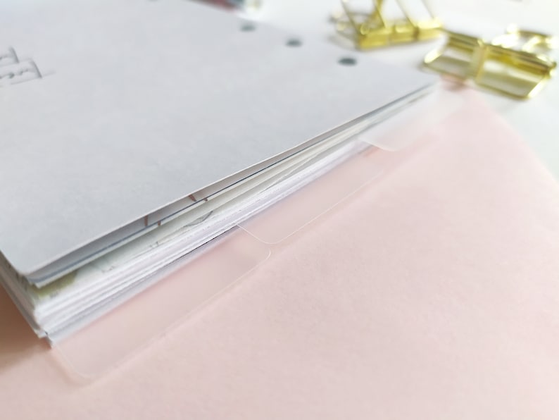 May include: A stack of white paper sheets with punched holes along the left edge, featuring translucent plastic index tabs extending from the right side. Two gold-toned metal binder clips are positioned near the top right corner of the paper stack.