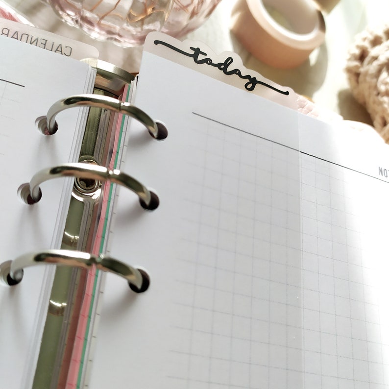 May include: An open planner with a grid-lined page and a clear divider labelled "today" in black script. The planner has silver metal rings and a colourful edge. Stationery items are visible in the background.