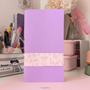 May include: A rectangular handmade notebook with a solid light purple cover. A light pink paper band wraps around the center, featuring a white botanical line art pattern and the text: handmade notebook, 60 sheets, versusprints.