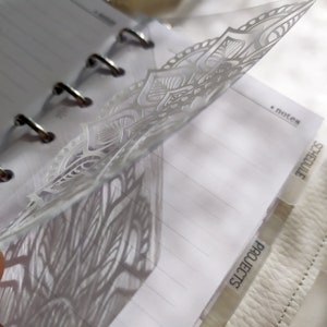 May include: A translucent plastic planner divider featuring a white mandala pattern. The divider is inserted into a ring-bound notebook with lined paper. Visible tabs on the side are labeled with the text "notes", "schedule", and "projects".