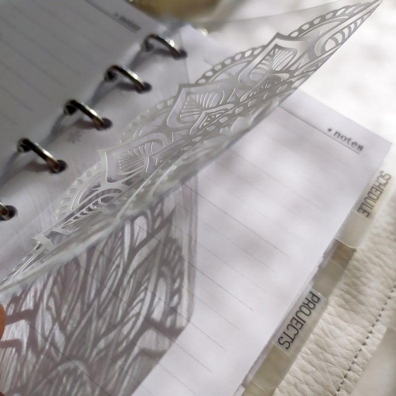 May include: A translucent plastic planner divider featuring a white mandala pattern. The divider is inserted into a ring-bound notebook with lined paper. Visible tabs on the side are labeled with the text "notes", "schedule", and "projects".