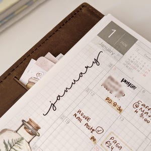 Può includere: Primo piano di un'agenda aperta sul mese di gennaio. La copertina in pelle marrone è visibile, insieme alle pagine a griglia. La parola "gennaio" è scritta in corsivo, con adesivi e note sul calendario.