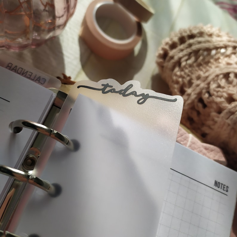 May include: A planner with a clear, frosted divider labelled "today" in black script. The planner is open, revealing lined pages and a metal binder. A roll of peach-coloured washi tape and a decorative glass jar are in the background.