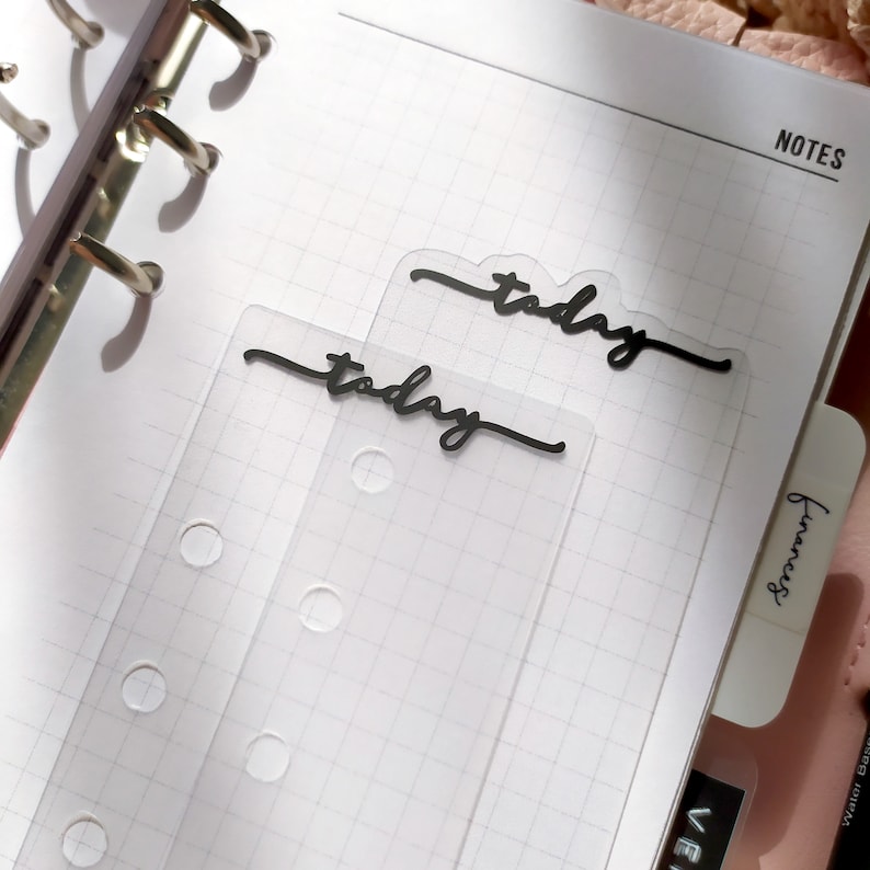 May include: Two transparent planner dividers, each with the word "today" in black script, rest on a grid-lined notebook page. The notebook is open, with the word "notes" printed at the top. The dividers have pre-punched holes.