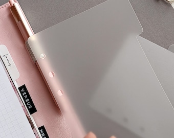 Könnte beinhalten: Ein hellrosa Planer-Ringbuch mit einem goldenen Stift und halbtransparenten, mattierten Kunststoff-Trennblättern. Die sichtbaren schwarzen Registerkarten tragen die Aufschriften "FINANCES", "VERSUS" und "MRS. L". Das Ringbuch enthält kariertes Papier und eine goldene Metallklemme.