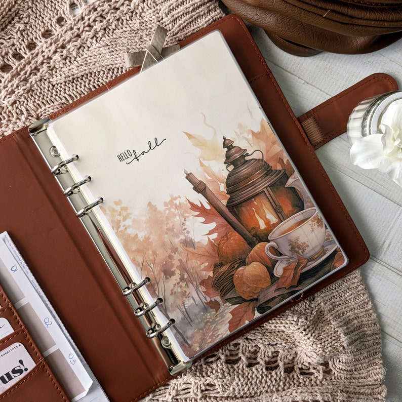 K&ouml;nnte beinhalten: Ein brauner Leder-Ringbuchplaner mit einer dekorativen Einlage mit dem Text Hello Fall. Die Illustration zeigt eine leuchtende Laterne, eine Teetasse und K&uuml;rbisse inmitten von Herbstbl&auml;ttern. Der Planer hat eine silberne Sechs-Ring-Mechanik.