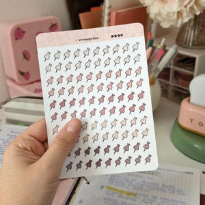 Peut inclure: Une planche d'autocollants rectangulaire présentant une grille de petites illustrations de punaises dans diverses nuances de rose, beige et marron. L'en-tête supérieur est rose clair avec le texte "versusprints" et des icônes de réseaux sociaux.