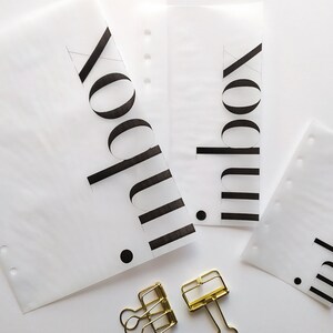 May include: Three translucent white planner inserts of varying sizes, each featuring the word inbox printed in bold black sans-serif font with a small black dot. Two gold-toned metal binder clips are positioned below the paper sheets.