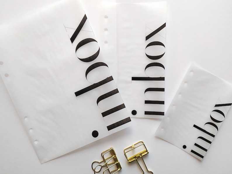 May include: Three translucent white planner inserts of varying sizes, each featuring the word inbox printed in bold black sans-serif font with a small black dot. Two gold-toned metal binder clips are positioned below the paper sheets.