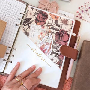 May include: A brown leather ring-bound planner featuring a white pocket with the word stickers printed in gold script. The planner contains floral patterned paper, a small card with the text find beauty every day, and a sticker of a person reading.