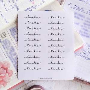 以下が含まれることがあります： 白い紙に「タスク」という言葉が12回、筆記体で書かれています。紙は、ピンクの花とノートが置かれたテーブルの上にあります。