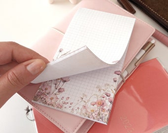 Puede incluir: Un bloc de notas rectangular con un patrón de cuadrícula blanca y una ilustración floral de flores silvestres rosas y marrones en el borde inferior. El bloc está sobre una funda de cuaderno rosa claro y una agenda color coral del año 2023.