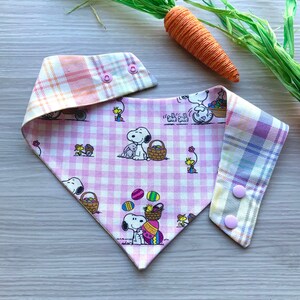 May include: Pink and white plaid dog bandana with a pink and white gingham pattern on the other side. The bandana features a cartoon image of Snoopy and Woodstock with Easter eggs. The bandana has two pink snap buttons.