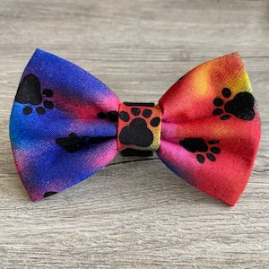 May include: A colorful bow tie with a rainbow tie-dye pattern and black paw prints. The bow tie is made of fabric and has a black elastic band.