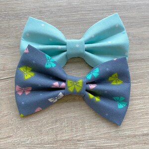 May include: Two fabric bow ties. One is light blue with white polka dots. The other is dark blue with a pattern of pink, green, and yellow bows.