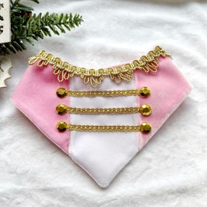 May include: A pink and white pet bandana with gold trim and button details. The bandana has a velvet texture and a decorative gold lace trim along the top edge. Three gold button accents are centered on the front.