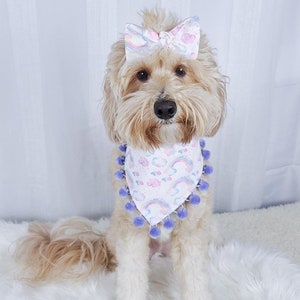 May include: A light brown dog wearing a white bow with a rainbow and cloud pattern. The dog also wears a matching bandana with purple pom-poms and a rainbow and cloud pattern. The dog is sitting on a white fluffy surface.