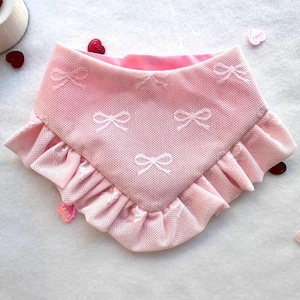 May include: A pink pet bandana with a bow pattern. The bandana has a ruffled edge and a soft pink lining. White bows are embroidered across the front of the bandana. The bandana is designed to be worn around a pet's neck.