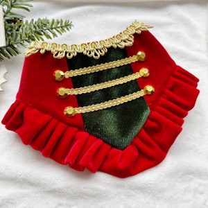 May include: Red velvet dog collar with a green velvet panel, gold trim, and gold buttons. The collar has a ruffled edge.