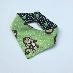 May include: A green and black baby bib with a monkey print. The bib is reversible, with a green side featuring white polka dots and cartoon monkeys, and a black side with colorful polka dots. It fastens with two green snaps.
