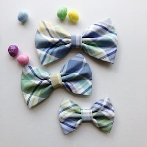 May include: Three plaid bow ties in different sizes. The largest bow tie is blue, green, and white. The medium bow tie is green, blue, and white. The smallest bow tie is blue, green, and white.