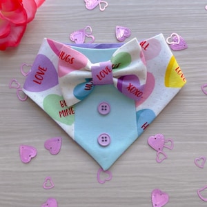 May include: A light blue dog bandana with a white background and a pattern of candy hearts with phrases like "Love", "Hugs", and "Be Mine". The bandana has a matching bow tie with a white background and a pattern of candy hearts. The bandana is on a light wood surface with pink heart-shaped confetti.