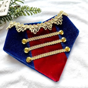 Puede incluir: Un pañuelo para mascotas de terciopelo azul y rojo con ribetes dorados y botones decorativos. El pañuelo tiene un diseño de estilo militar con adornos dorados en los bordes y tres filas de botones dorados. El fondo es de tela blanca suave.