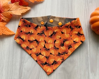 Op de afbeelding: Een omkeerbare bandana met een bruin en zwart ruitpatroon aan de ene kant en een pompoenprint aan de andere kant. De bandana heeft drie gele drukknopen.