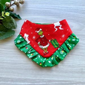 May include: Red and green Christmas dog bandana with a white snowflake pattern and a Grinch character wearing a Santa suit. The bandana has a ruffled edge.