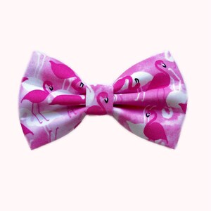 May include: Pink bow tie with a flamingo print. The bow tie is made of fabric and has a white background with pink flamingos.