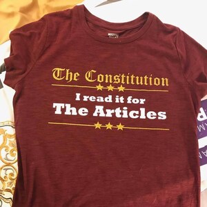 May include: Burgundy t-shirt with a white graphic that reads "The Constitution I read it for The Articles" with gold stars.