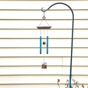 May include: A hanging wind chime with a dark metal frame and a small house image at the bottom. The chime has blue rectangular rods and a brown sphere in the center. The wind chime is hanging from a black metal hook.