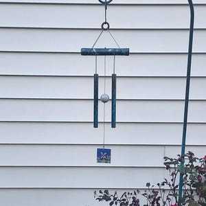 May include: A handcrafted wind chime with a dark blue and green finish. The chime features a horizontal bar, vertical tubes, a central sphere, and a small square tile with a floral design. The wind chime is suspended from a metal hook.