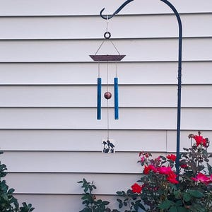 May include: A wind chime featuring a dark wooden top bar, two blue cylindrical tubes, a central copper-colored sphere, and a square white wind catcher at the bottom decorated with a black and white graphic illustration of two figures.
