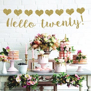 May include: A table setting with a pink and gold theme. There are several cakes and desserts on the table, including a cake with gold drip icing, a tower of macarons, and a layered cake. The table is decorated with flowers and greenery. The banner above the table says "Hello twenty!"
