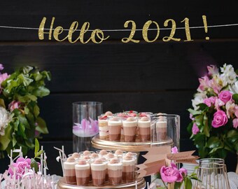 May include: A dessert table decorated with pink and white flowers, featuring a variety of desserts, including chocolate mousse, cake, and fruit. A banner with the text "Hello 2021!" hangs above the table.