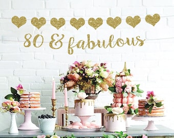 May include: A table setting for an 80th birthday party with a gold glitter banner that says "80 & fabulous". The table is decorated with pink and white cakes, flowers, and candles.