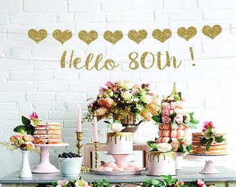 May include: A table setting for a birthday party with a gold glitter banner that says "Hello 80th!"  The table is decorated with flowers, candles, and cakes.