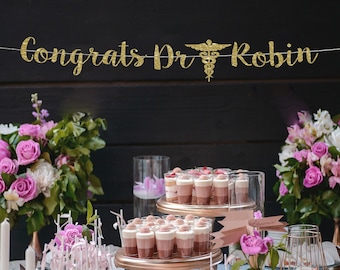 Peut inclure: Une bannière dorée à paillettes avec le texte "Congrats Dr Robin" et un symbole de caducée. La bannière est suspendue au-dessus d'une table avec des fleurs roses et des desserts dans des bocaux en verre.