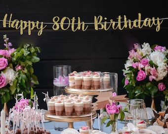 May include: A gold glitter banner with the text "Happy 80th Birthday" hanging above a dessert table with pink flowers, cupcakes, and small desserts in glass jars.