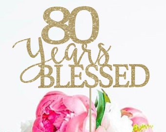 May include: Gold glitter cake topper with the text "80 Years Blessed" on a white cake with pink flowers.
