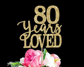 May include: Gold glitter cake topper with the text "80 years loved" on a white cake with pink flowers.