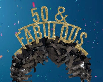 May include: A black and gold 50th birthday headband. The headband features the words "50 & FABULOUS" in gold glitter, above a fringe of black paper. The headband is on a blue background with confetti.