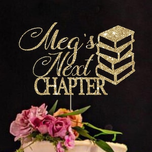 May include: A gold glitter cake topper featuring the text Meg's Next Chapter and a stack of three books, positioned above a white frosted cake decorated with pink and orange flowers on a decorative metallic stand with a scalloped edge.