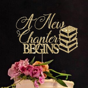 May include: A gold glitter cake topper featuring the text A New Chapter BEGINS and a stack of three books. The topper is positioned above a white frosted cake decorated with pink and orange flowers on a decorative gold cake stand.