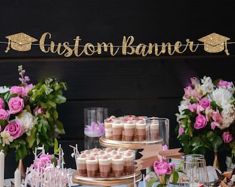 May include: Gold glitter graduation cap banner with the text "Custom Banner". A table decorated with pink flowers and small desserts in clear glass cups.