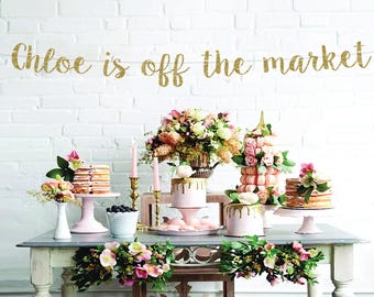May include: A bridal shower dessert table with a gold glitter banner that reads "Chloe is off the market". The table is decorated with pink and white flowers, and there are several cakes and cupcakes on display.