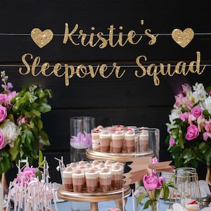 May include: A gold glitter banner with the text Kristie's Sleepover Squad, featuring a heart icon on each side of the name. The banner is suspended against a dark background above a table with floral arrangements and small dessert cups.