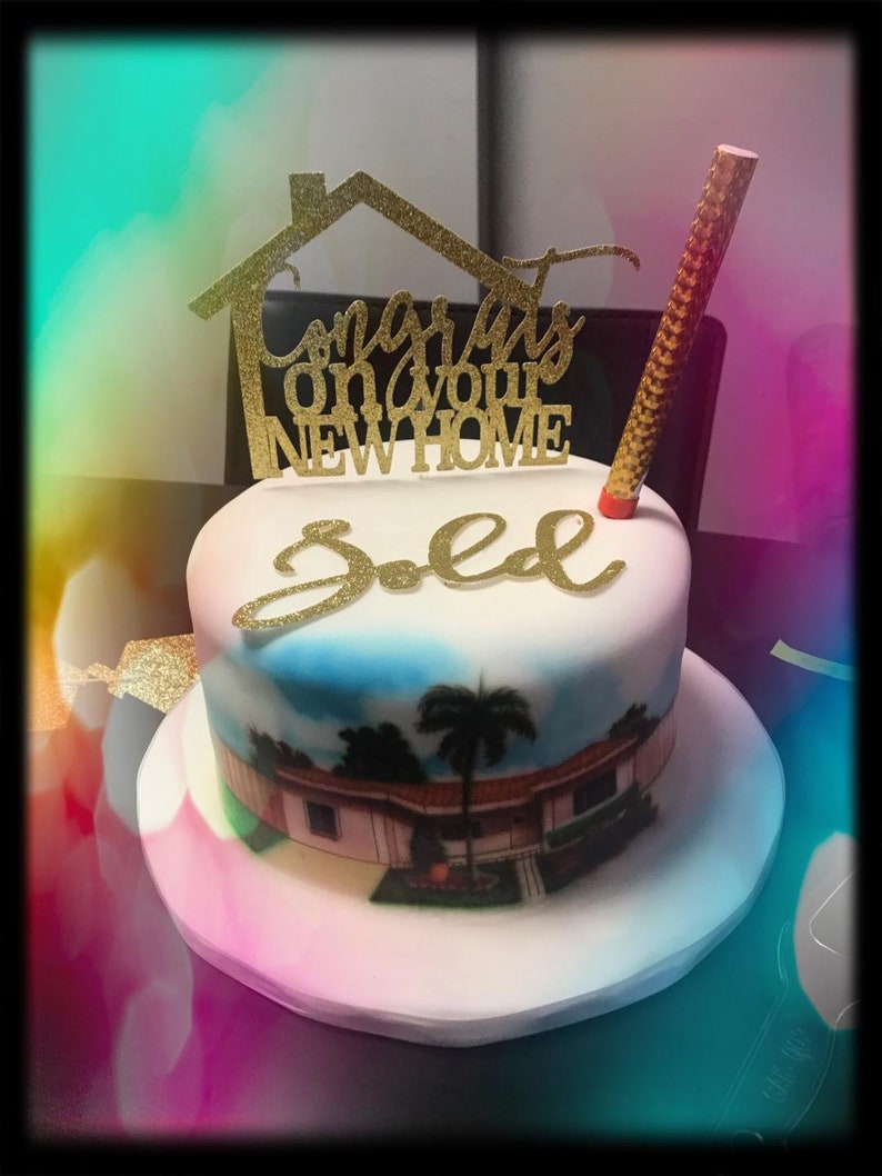 May include: A white cake with a blue and green image of a house with palm trees. The cake is decorated with a gold glitter topper that says "Congrats on your new home" and a gold glitter "Sold" sign.
